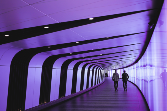 Two men walk through a purple tunnel with black stripes and a shiny wall on the right.