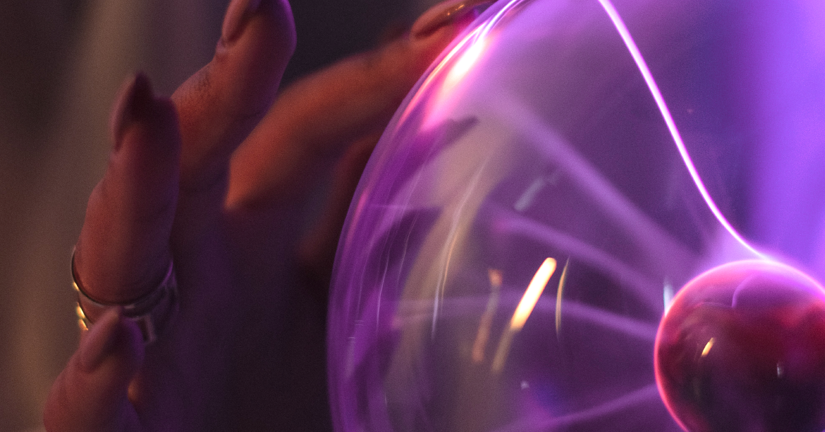A close-up of a woman's hand touching a glass tesla coil. She is wearing rings and nail polish.
