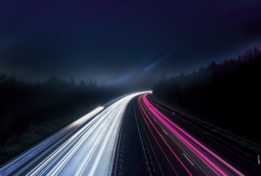 Stylized stop motion photo of a dark forest transected by a highway with streaks of light depicted as head and tail lights.