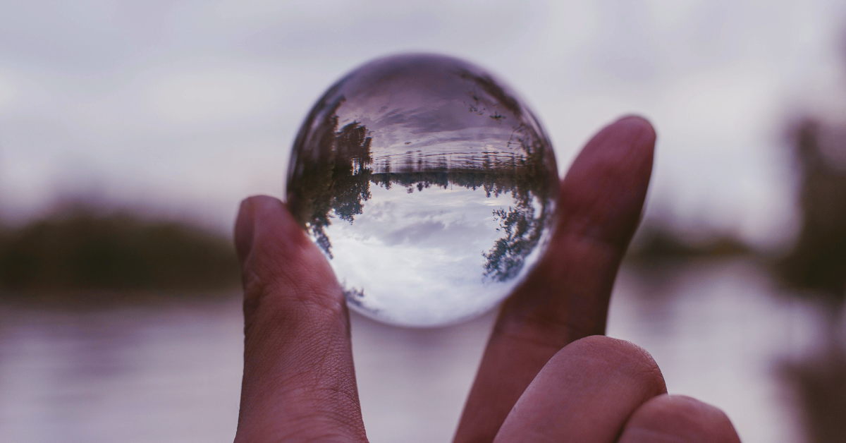 A thumb and forefinger hold a glass orb that shows sky, water and trees upside down.